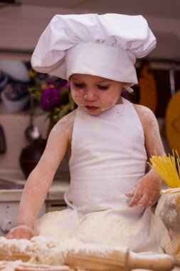 Beyaz bir aşçı elbisesi içinde bir çocuk masada oturuyor. Masanın üzerinde süt, un ve oklava var. Bir çocuk hamur yoğurur.
