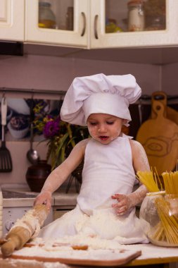 Beyaz bir aşçı elbisesi içinde bir çocuk masada oturuyor. Masanın üzerinde süt, un ve oklava var. Bir çocuk hamur yoğurur.