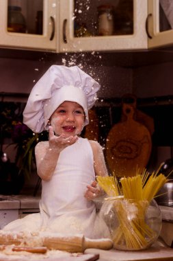 Un ile oynanan aşçı kostümü giymiş küçük bir çocuk. Masanın yanında hamur, merdane ve un var. Çocuk yukarı bakar ve güler. Evin içinde çok olumlu bir fotoğraf var.