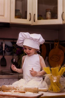 Beyaz bir aşçı elbisesi içinde bir çocuk masada oturuyor. Masanın üzerinde süt, un ve oklava var. Bir çocuk hamur yoğurur.