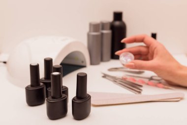 Manikür ustasının elinde manikür dekoru var. Yakınlarda jöle cilası, manikür aletleri ve ultraviyole lamba var.