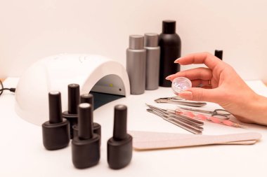 Manikür ustasının elinde manikür dekoru var. Yakınlarda jöle cilası, manikür aletleri ve ultraviyole lamba var.