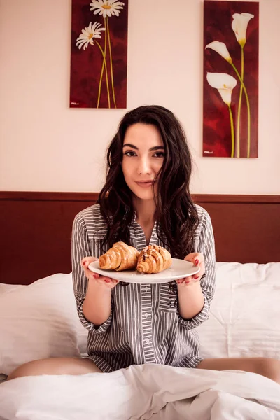 A beautiful young woman sits in a bed and holds croissants in her hands. A girl with dark hair. She has a slight smile on her face. The girl rests. Two paintings hang on the wall