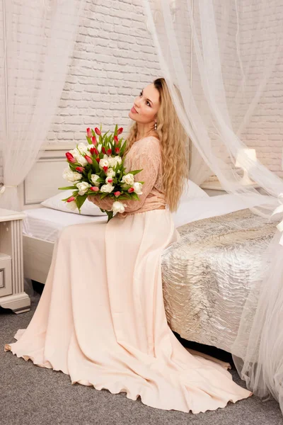 Beautiful young blonde in a light dress with a bouquet of tulips in her hands. She sits on a bed in a hotel room. Her gaze is directed upwards, she smiles.