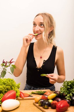 Tahta masada taze sebzeler, biberler, domatesler, patlıcanlar, marul, salatalık, maydanoz var. Mutfakta genç bir kadın salatalık yiyor. Vejetaryen Günü. Yüksek kalite fotoğraf