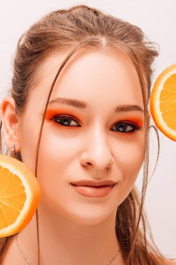 Kız yüzünün yanında iki tane sulu portakal tutuyor. Parlak makyajlı ve portakallı güzel bir kızın portresi