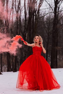 Gür kırmızı elbiseli güzel genç bir sarışın karlı bir ormanda duruyor. Kışın elinde kırmızı dumanla kırmızı elbiseli güzel sarışın kadın.