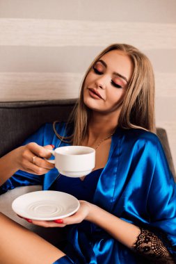 Bir kadın elinde beyaz bir fincan kahve ve bir fincan çay tabağı tutuyor. Gözlerini kapadı ve sabah kahvesinin tadını çıkardı. Mavi ipek bornoz. - Günaydın. Dawn.