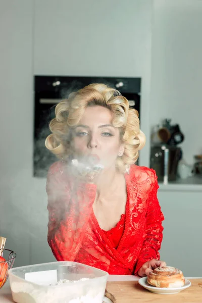 A beautiful woman in a red robe holds flour on her hand and blows air on it. The image of a happy housewife in a red lace robe. Large curls on the hair. Proper nutrition, diet, cooking in the kitchen