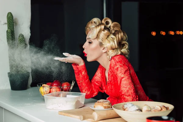 A beautiful woman in a red robe holds flour on her hand and blows air on it. The image of a happy housewife in a red lace robe. Large curls on the hair. Proper nutrition, diet, cooking in the kitchen