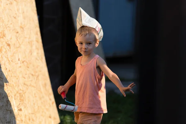 Küçük sarışın çocuk inşaatçılık oynuyor. Tişörtlü, şortlu ve garnizon şapkalı çocuk duvarları boyuyor. — Stok fotoğraf