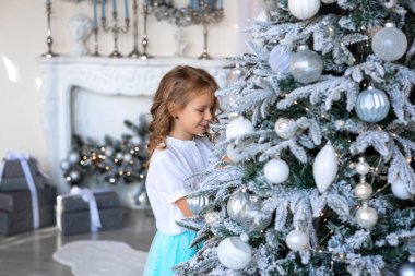 Sarışın bir kız oyuncağını asmak için Noel ağacına uzanır.