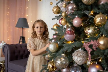 Sarışın bir kız oyuncağını asmak için Noel ağacına uzanır.