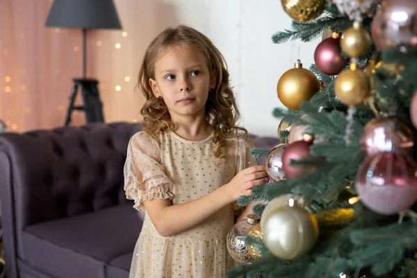 Sarışın bir kız oyuncağını asmak için Noel ağacına uzanır.