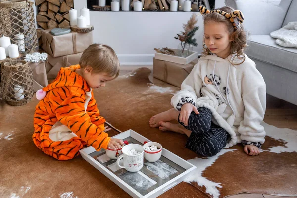 Kigurumi pijamalı çocuklar yere oturup kakao ve kurabiye içerler.