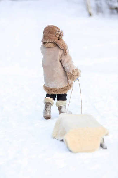 Un bambino porta una slitta da una corda su una strada innevata. Un ragazzo in abiti popolari invernali — Foto Stock