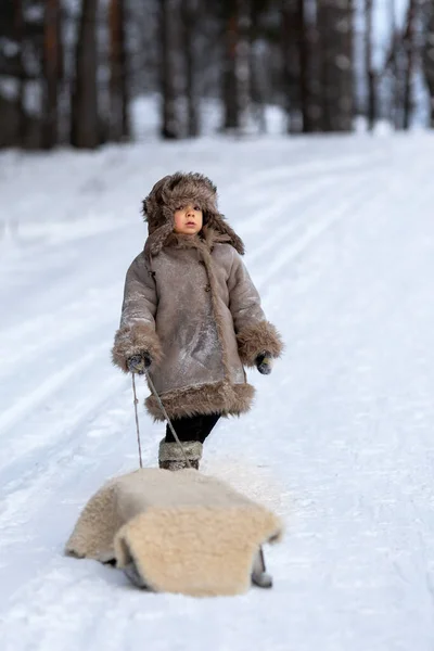 Ребенок несет сани на веревке по заснеженной дороге. Мальчик в зимней народной одежде — стоковое фото