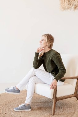 Minimum ışık ve havadar iç tasarım. Beyaz kotlu ve yeşil kazaklı çekici genç bir kadın açık ve havadar iç kısımda sandalyede oturuyor.