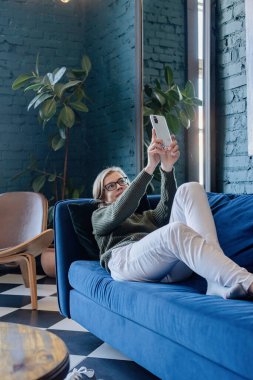 Yeşil kazaklı ve beyaz kotlu genç beyaz kadın şık mavi oturma odasında kanepede uzanıyor ve cep telefonuyla selfie çekiyor.