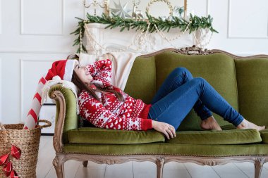 Mutlu Noeller ve mutlu yıllar. Noel Baba şapkalı genç esmer kadın yeşil koltukta tek başına Noel için dekore edilmiş oturma odasında yatıyor.