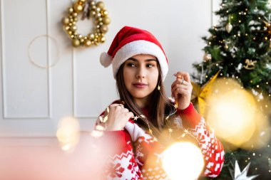 Noel Baba şapkalı ve kazaklı genç bir kadın elinde güzel noel ışıkları tutuyor.
