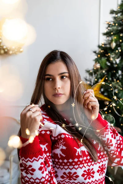 Noel Baba şapkalı ve kazaklı genç bir kadın elinde güzel noel ışıkları tutuyor.