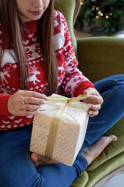 Tatil hediyelerini paketliyorum. Noel hediyesi paketi. Kırmızı kazaklı genç bir kadın kanepeye oturmuş hediyesini paketliyor.