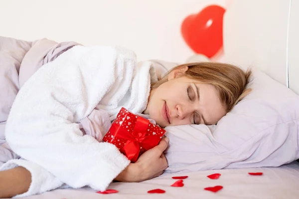 Mujer rubia joven acostada en la cama con regalo de vacaciones y globos rojos — Foto de Stock