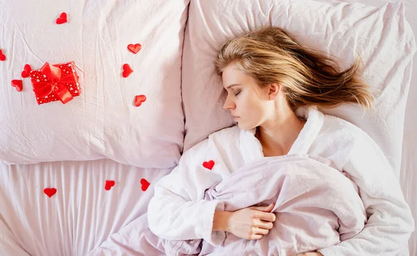 Mujer rubia joven acostada en la cama con regalo de vacaciones y confeti rojo — Foto de Stock