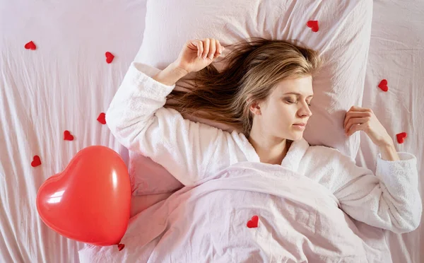 Mujer rubia joven acostada en la cama con regalo de vacaciones y confeti rojo — Foto de Stock