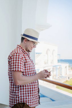 Tatilde. Yazlık şapkalı yakışıklı beyaz adam otelin balkonunda duruyor, cep telefonu kullanıyor.