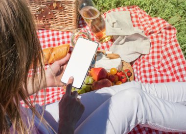 back view of unrecognizable woman in white pants outside having picnic and using smartphone taking photo. Summer fun and leisure. view from behind