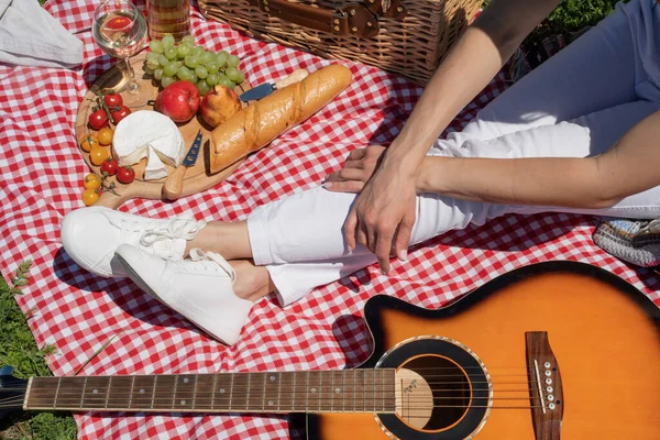 Top view of unrecognizable young woman in white pants outside having picnic, eating and playing guitar. Summer fun and leisure