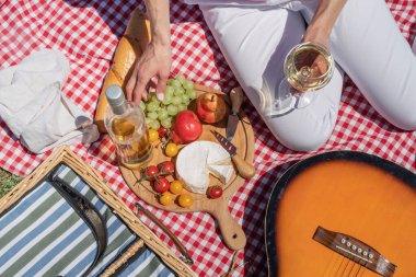 Top view of unrecognizable young woman in white pants outside having picnic, eating and playing guitar. Summer fun and leisure