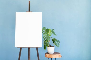Wooden easel with blank canvas with monstera plant on light blue wall background for mockup design. Space for text