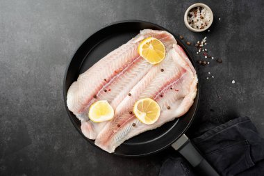 Fresh raw white fish fillet Pangasius with spices and lemon in frying pan on dark stone background