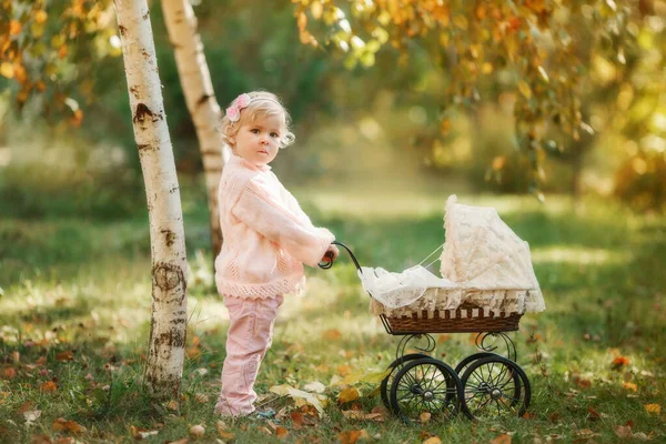 Красива блондинка маленька дівчинка на відкритому повітрі, зупиняючись біля дитячої коляски для ляльок і посміхаючись . — стокове фото