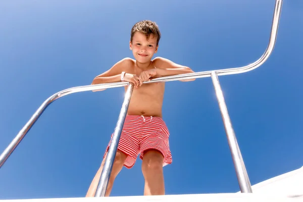 Bir yat güvertesinde duran ve güneşli bir günde batacak olan genç bir çocuk. Çocuk şort giyiyor, alttan bakıyor..