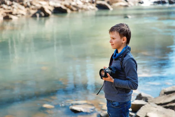 Fotoğrafçı çocuk bir dağ nehrinin kenarında kayanın üzerinde duruyor ve bir şeye bakıyor. Çocuk güneşli bir günde güzel mavi suyun tadını çıkarıyor ve gülümsüyor. Çocuk fotoğraf makinesi tutuyor..