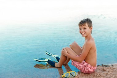 Plajda tropikal yaz tatilinde tatlı bir çocuk. Çocuk deniz kenarındaki kumda oturuyor, mayo, palet giyiyor ve kameraya bakıyor. Sahilde yaz zamanı. 