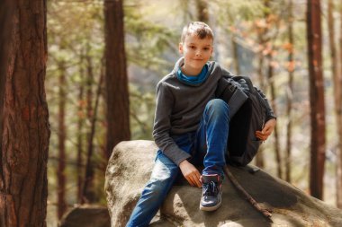 A boy sits on a large stone in nature. Kid is smiling and looking at camera, backpack is near the person. Child has a good mood during autumn sunny day.