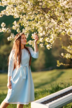  Portrait of beautiful happy woman in blue dress with long hair staying near blooming tree with synthesizer piano.