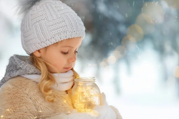 Cute little girl playing on snowy winter day in the park. Looking at the garland. Waiting for miracle.
