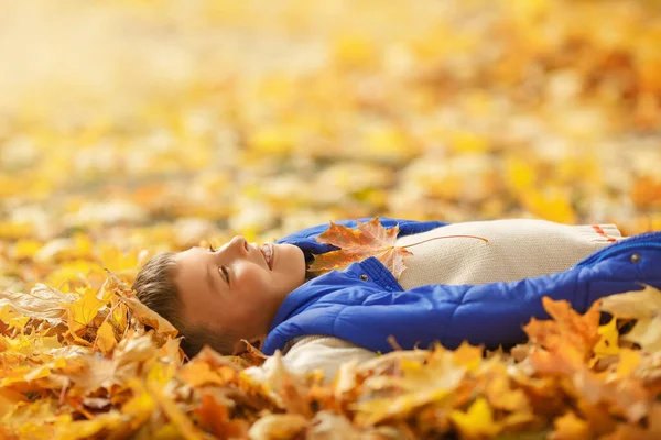 Happy boy in sweater and blue vest laying on autumn leaves and smiling. Autumn holiday time.
