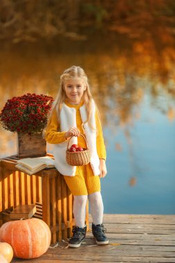 Kız sonbahar günbatımında, bir ahşap iskelede nehrin kenarında duruyor. Balkabağı ve çiçekler çocuğun yanında..