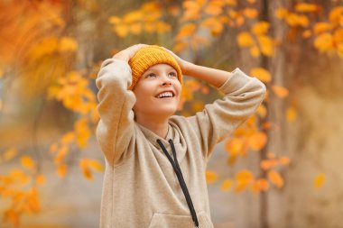 Sonbahar günü bej kazaklı ve sarı şapkalı çocuk sarı ağacın önünde duruyor. İki elini de başının üstüne kaldır. Çocuk ayakta duruyor ve güzel bir ağacın yanında gülümsüyor. Temiz hava..