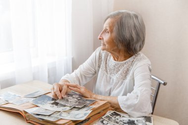 Mature woman watching black and white old photo album at home.Person looking at her own photo was taken in 1963, 1965. She has got smile while remembering how young she was and looking at the window. clipart