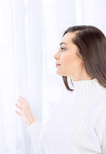 a girl in white stands and looks out the window