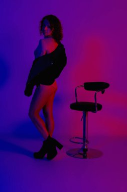Stüdyodaki neon ışıklarda şık kıyafetler giymiş bir manken. Deri ceketli seksi bir kadının stüdyo portresi, kırmızı, mavi, mor morötesi bir gece kulübünde vücut ve botlar.