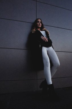 Akşamları karanlık bir duvara karşı poz veren seksi kız. Uzun siyah ceketli, beyaz kotlu, deri çantalı şık bir kız binanın duvarının yanında duruyor. Yürüyüşe çıkan güzel kız.